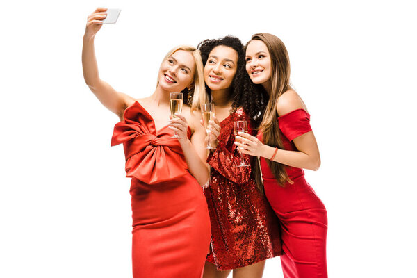 beautiful multiethnic girls in red dresses with champagne glasses taking selfie on smartphone isolated on white