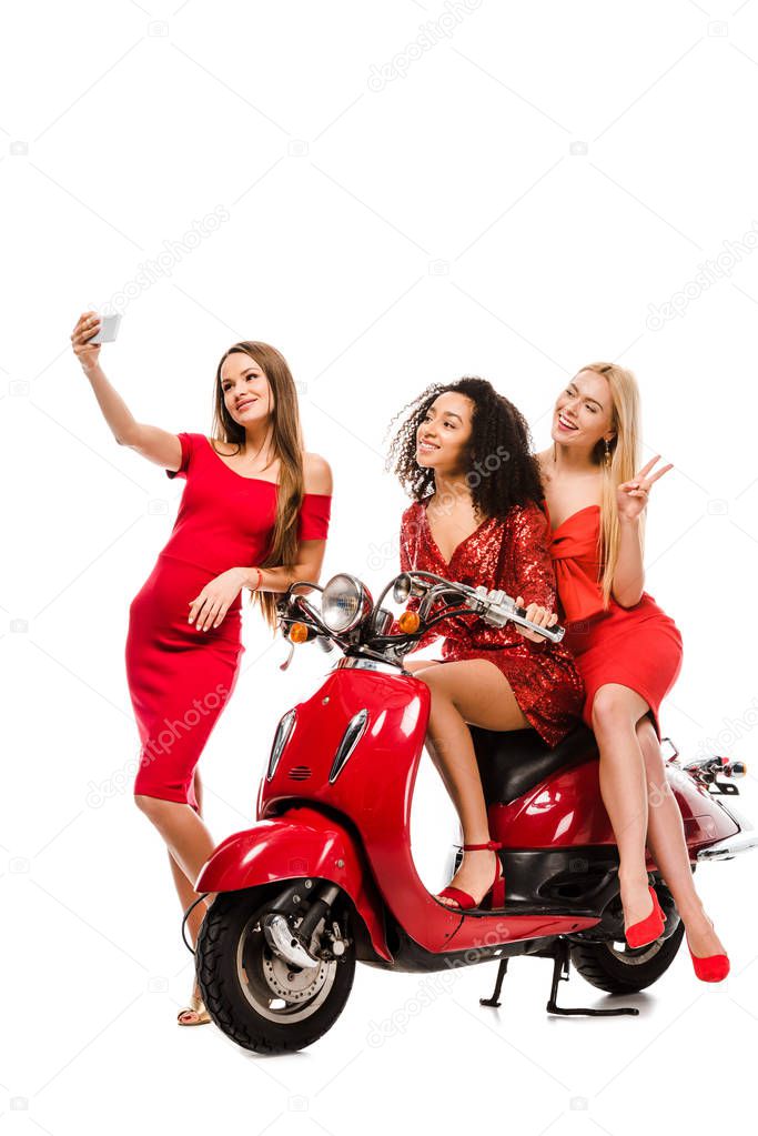beautiful multiethnic girls in red dresses on motor scooter taking selfie on smartphone isolated on white