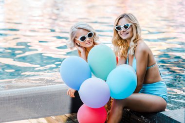 happy pin up girls in swimwear holding balloons while sitting at poolside