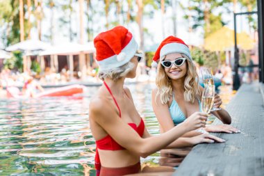 happy women in swimsuits and santa hats holding champagne glasses and sitting in swimming pool