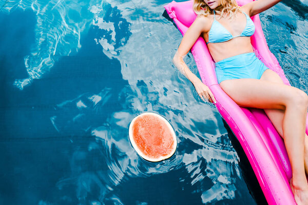 cropped view of girl in swimsuit resting on pink inflatable mattress in swimming pool with watermelon