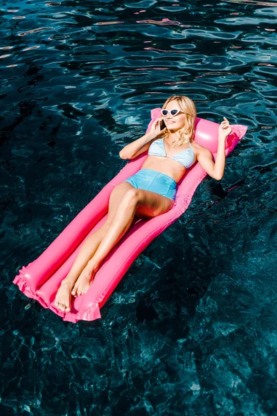 美丽的女孩在智能手机上交谈 而放松在粉红色的充气床垫在游泳池 — 图库照片