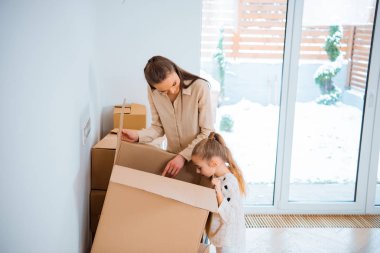 happy mother standing near cute daughter looking inside box in new home  clipart