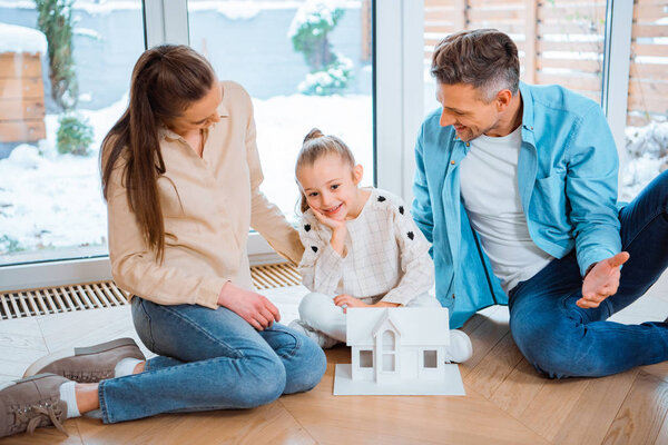 happy husband and wife looking at smiling daughter near house model while sitting on floor in new home