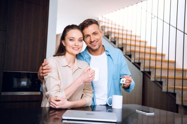 attractive woman holding cup of coffee and standing with cheerful husband holding house shaped key chain  clipart