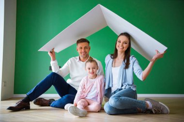happy husband and wife holding paper roof while sitting on floor near daughter clipart