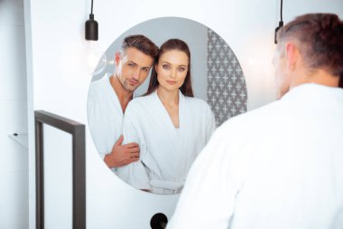 handsome husband hugging attractive wife in bathroom while looking at mirror clipart