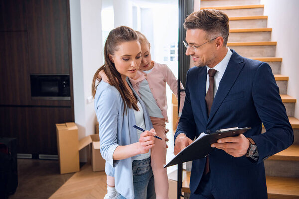 attractive woman holding in arms daughter and signing document near handsome broker holding clipboard 