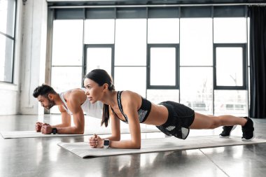 side view of athletic young couple in sportswear doing plank exercise on yoga mats in gym  clipart