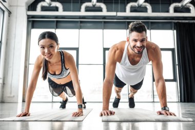 young couple doing push ups on yoga mats and smiling at camera in gym clipart