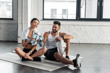 Genç çift yoga paspaslar üzerine oturan ve spor olarak çalışma sonra şişe su tutan spor giyim gülümseyen