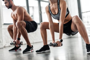 cropped shot of sporty young man and woman squatting with dumbbells in gym clipart