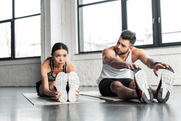 athletic young couple stretching legs and exercising on yoga mats in gym