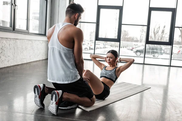 男教练员帮助年轻的女运动员在健身房做腹肌运动 — 图库照片