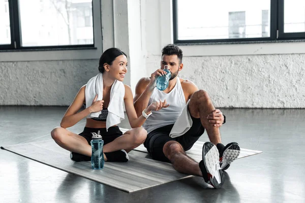 athletic young couple drinking water and resting on yoga mats after workout in gym