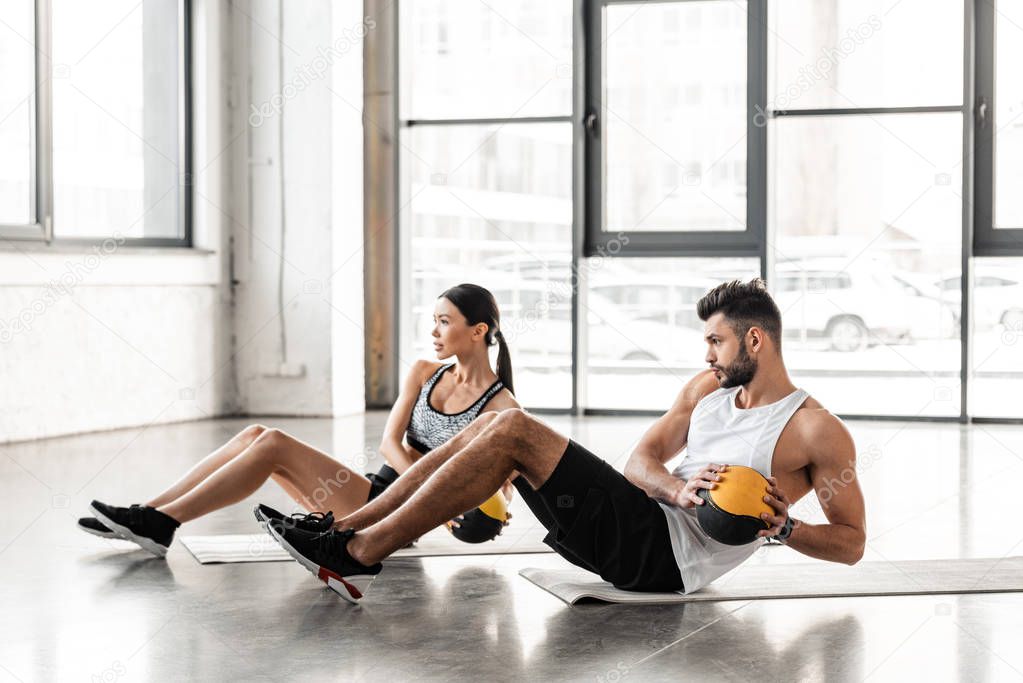 side view of athletic young couple in sportswear holding medicine balls and exercising on yoga mats in gym
