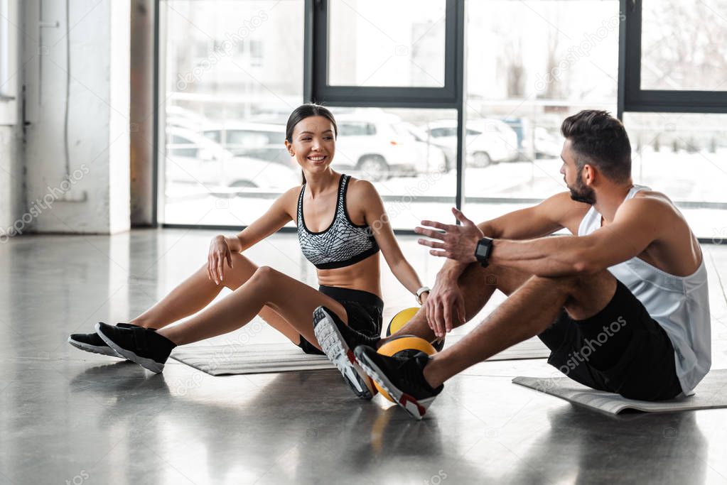 athletic young couple in sportswear sitting on yoga mats and talking in gym