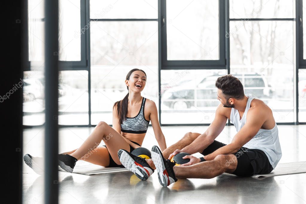 cheerful sporty young couple sitting on yoga mats with medicine balls and smiling each other in gym