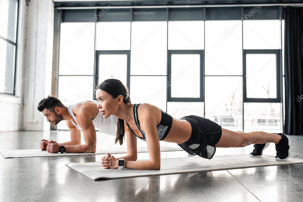 side view of athletic young couple in sportswear doing plank exercise on yoga mats in gym 