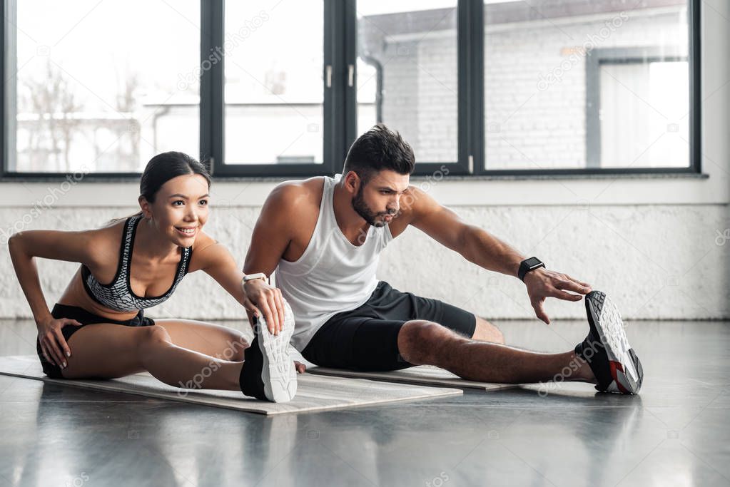 athletic young man and woman stretching legs on yoga mats in gym