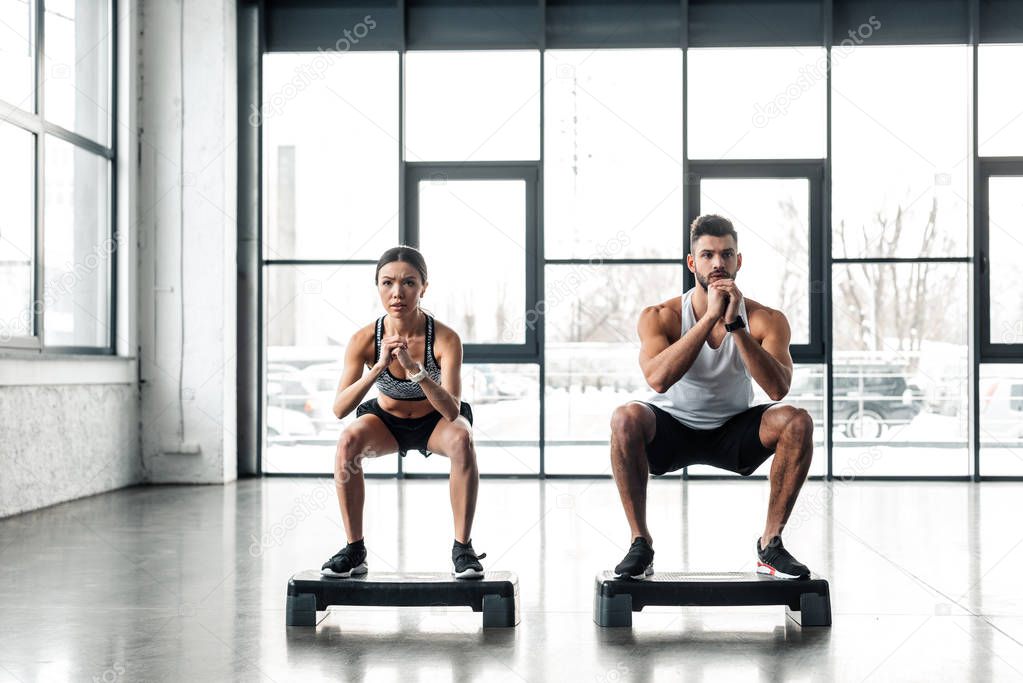 full length view of athletic young couple squatting on step platforms in gym