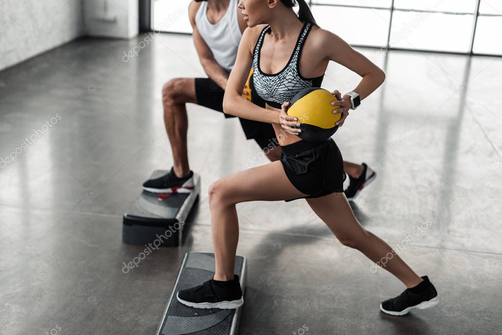cropped shot of sportive young couple holding medicine balls and exercising on step platforms in gym