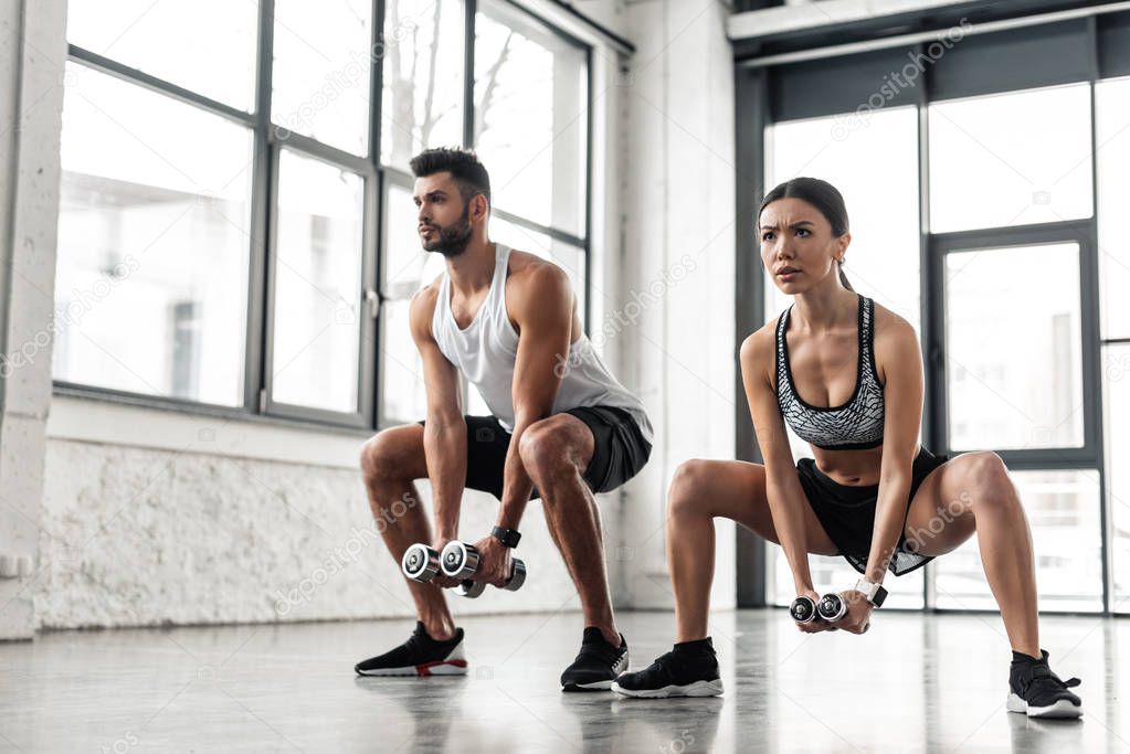 sporty young man and woman squatting with dumbbells in gym