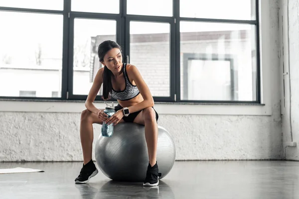 在健身房里 美丽的年轻女运动员坐在健康的球上 拿着一瓶水的全长视野 — 图库照片