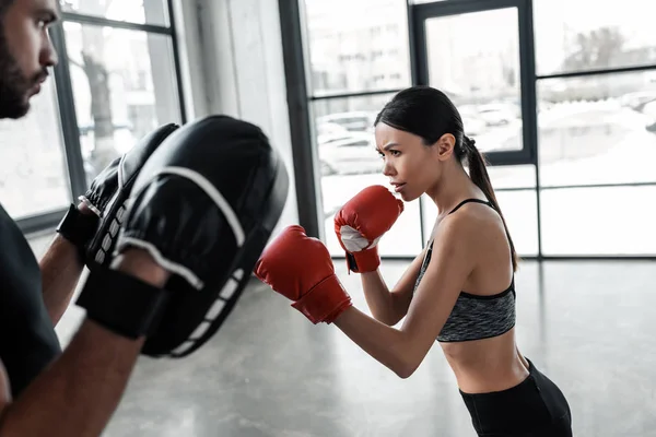 side view of concentrated young sportswoman in boxing gloves exercising with male trainer in gym