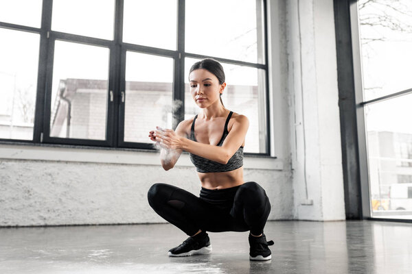 athletic young woman in sportswear crouching and applying talcum powder on hands in gym