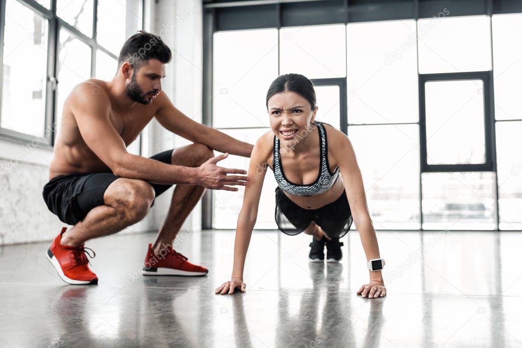 muscular shirtless male trainer helping young sportswoman doing push ups in gym