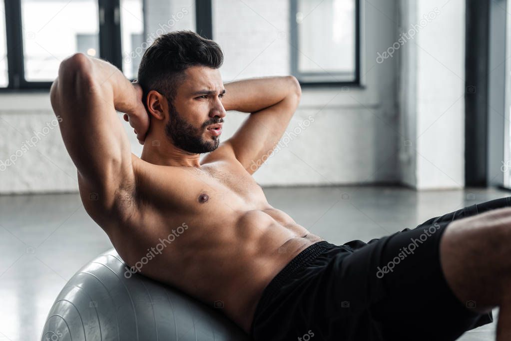 handsome young sportsman with bare chest doing abs exercise on fitness ball at gym