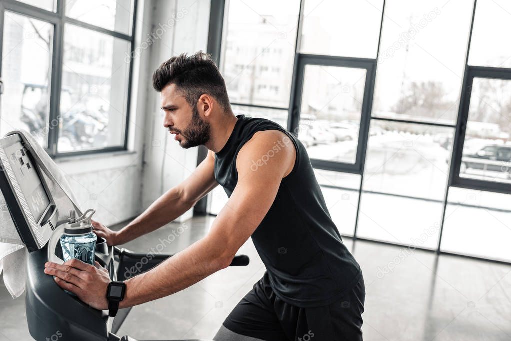 side view of athletic young man holding bottle of water and training on treadmill in gym