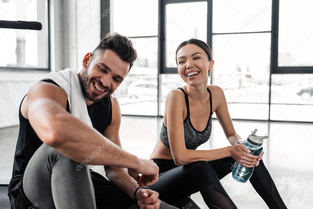 cheerful young couple laughing while sitting on treadmill and resting after workout in gym