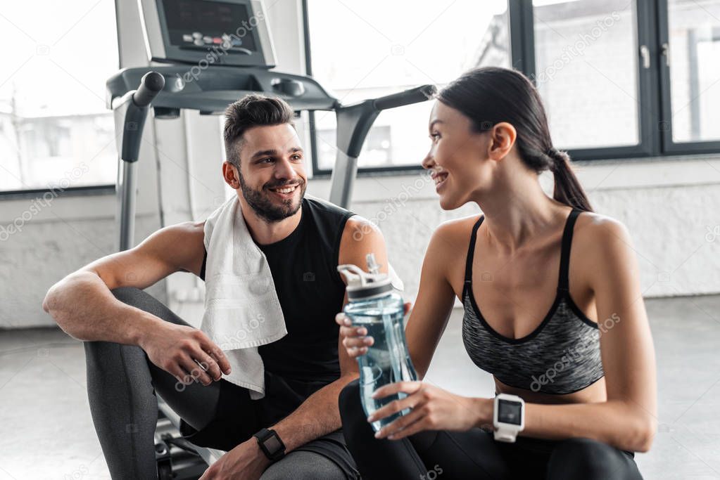 smiling young couple with towel and bottle of water resting together on treadmill in gym 