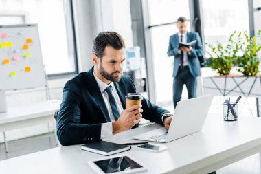 seçici odak yakışıklı işadamı kağıt bardak içki ile dizüstü bilgisayar kullanırken holding