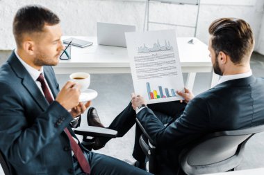 selective focus of businessman holding paper with graphs near coworker with cup of coffee clipart
