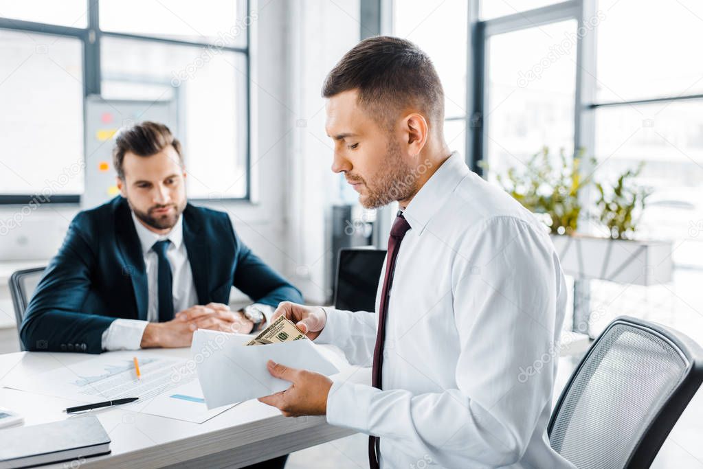 selective focus of businessman holding envelope with bribe near colleague in modern office 