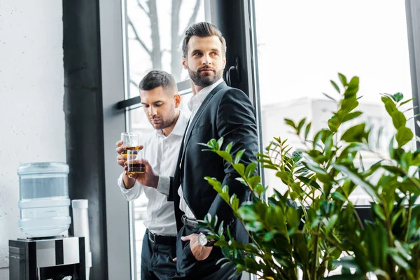 Biznesmen Stojąc Ręką Kieszeni Trzymając Szklankę Whisky Pobliżu Współpracownika — Zdjęcie stockowe