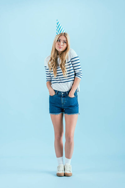 Long-haired girl in party hat posing with hands in pockets on blue background