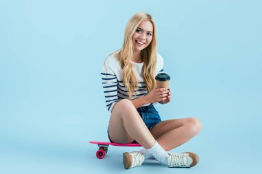 Mavi zemin üzerine kahve ile longboard üzerinde oturan güzel kız