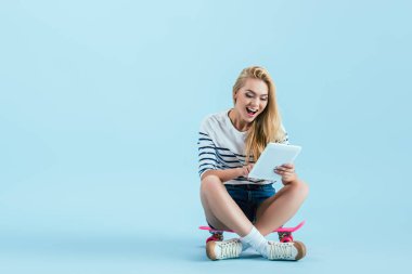 Heyecanlı kız mavi zemin üzerine longboard üzerinde otururken dijital tablet kullanma