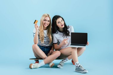 Kredi kartıyla longboard ve laptop ile mavi arka plan boş ekran üzerinde oturan güzel kızlar