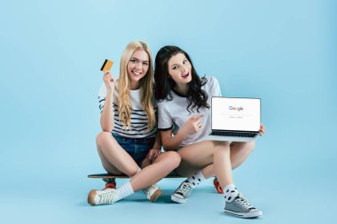 Longboard üzerinde oturan ve laptop ile google web sitesinde ekran ve kredi kartı mavi arka plan üzerinde tutarak çarpıcı kız