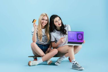 Kredi kartı ve online alışveriş sitesi olan dizüstü bilgisayar ekranında mavi arka plan üzerinde tutarak longboard neşeli kızlar