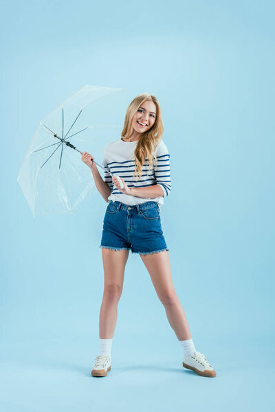 Smiling blonde woman in denim shorts holding umbrella on blue background