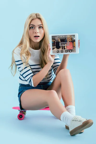 惊呆了的金发女孩坐在长板上 拿着数字平板电脑 在蓝色背景的屏幕上有在线预订应用 — 图库照片
