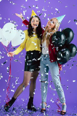 Happy girls in party hats posing with balloons under sparkle confetti on purple background clipart