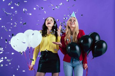 Happy girls with black and white air balloons posing under confetti on purple background clipart