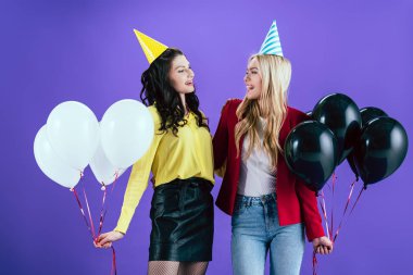 Cheerful girls in party hats holding balloons and looking at each other on purple background clipart